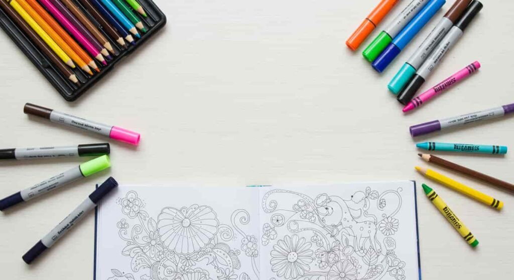 A flat lay of various coloring mediums including colored pencils, markers, and crayons, arranged around an open coloring book showing both intricate adult designs and simple kids' drawings, symbolizing choice and versatility for all ages